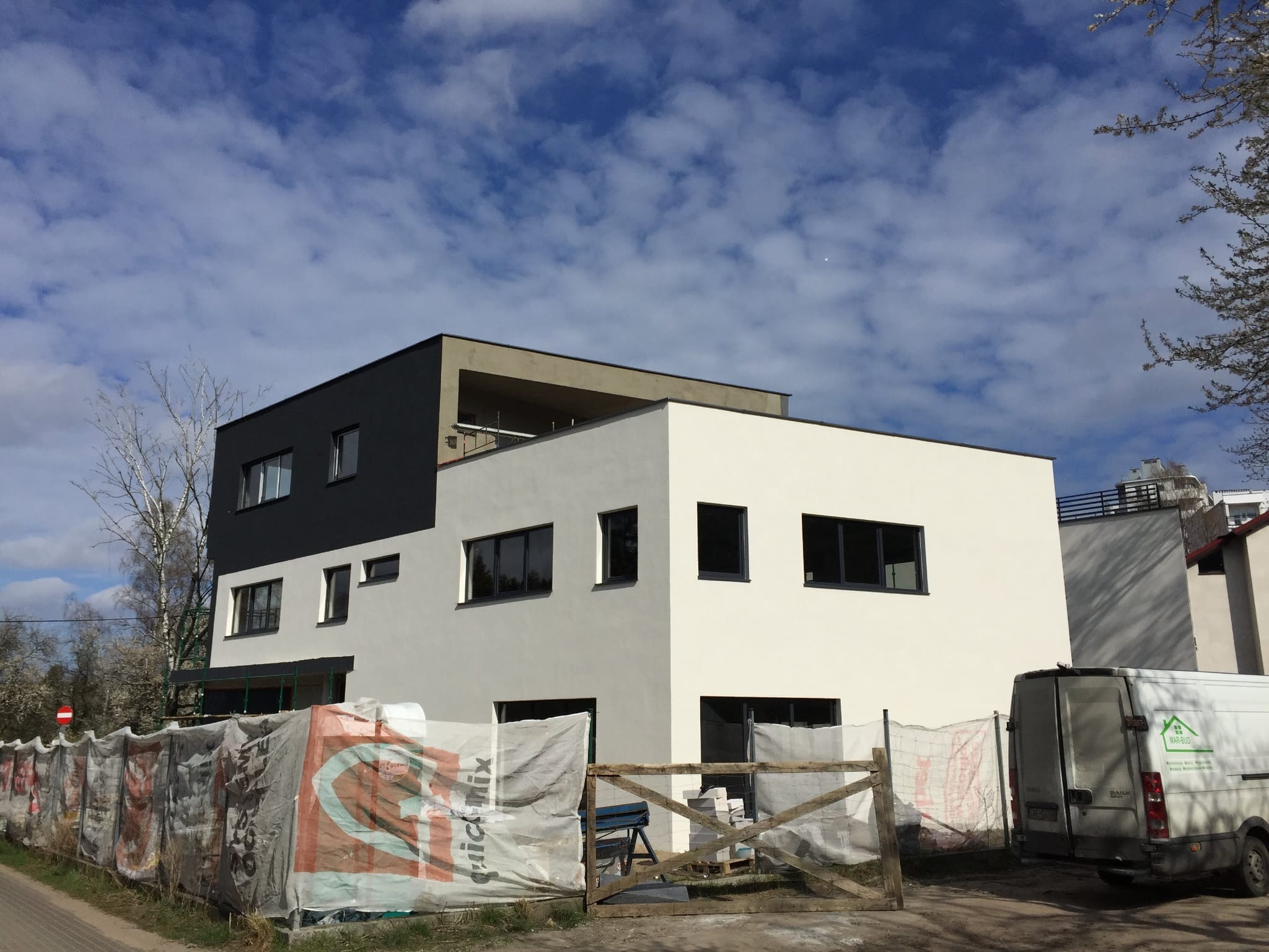 Nowoczesny dwukolorowy dom z płaskim dachem na etapie wykończenia elewacji, widoczny zza tymczasowego ogrodzenia i samochodu dostawczego z logo firmy budowlanej.