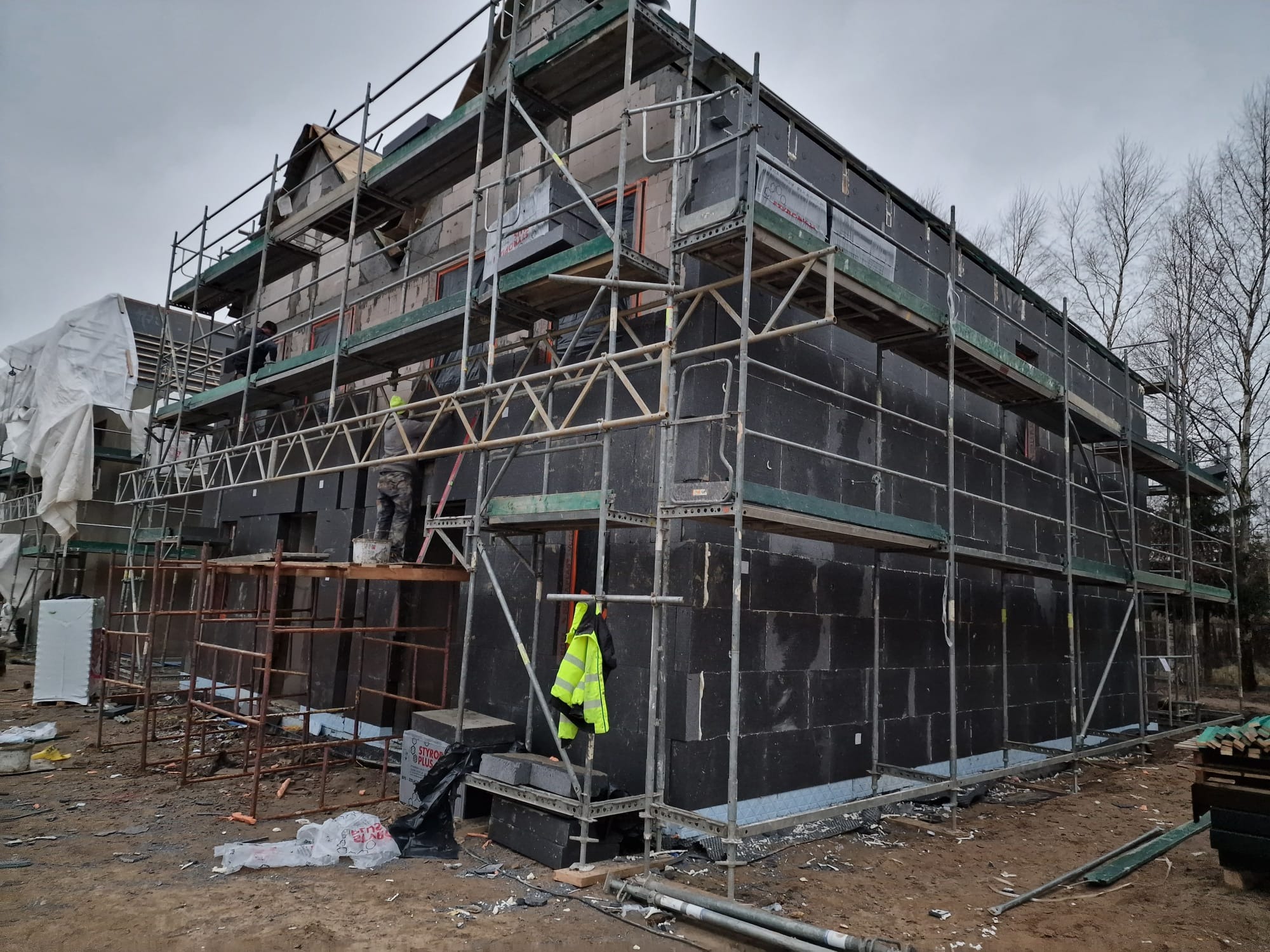 Budowa domu jednorodzinnego: elewacja w trakcie ocieplania styropianem grafitowym, widoczne rusztowania i pracownik z wiadrem, teren budowy z materiałami.