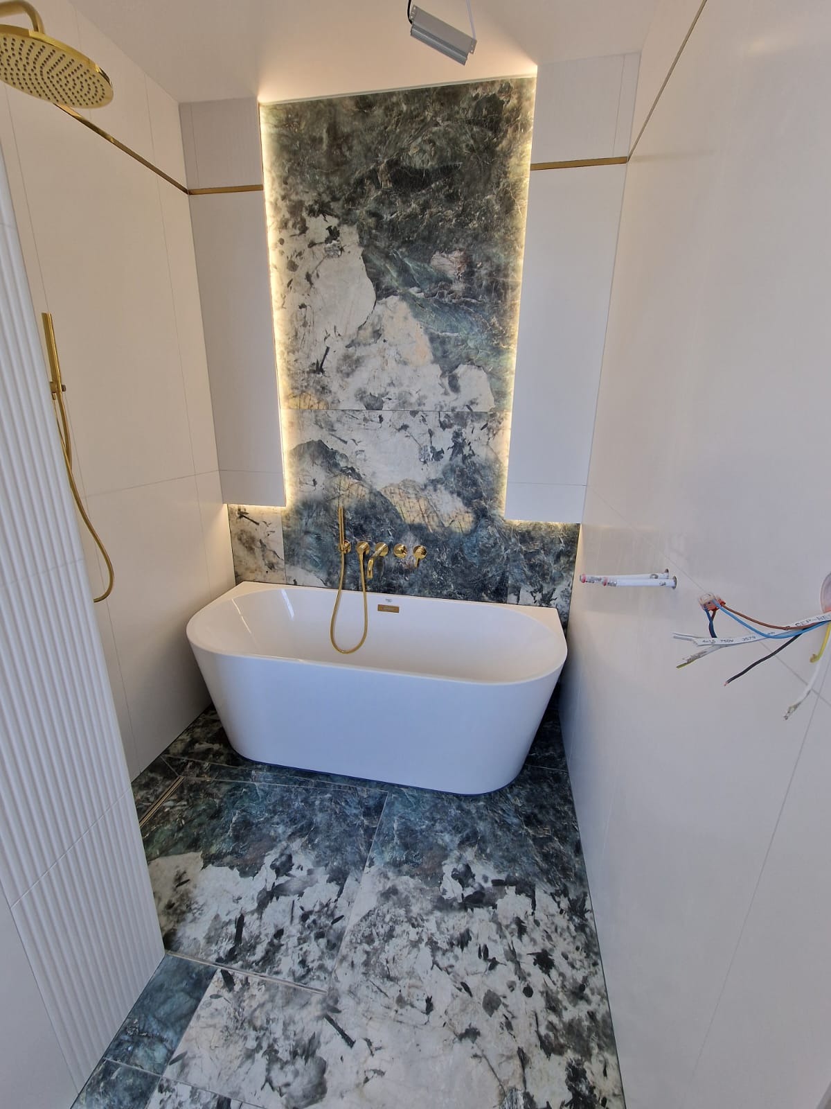 Nowoczesna łazienka z białą wanną wolnostojącą, złotą armaturą i marmurową ścianą z podświetleniem LED, widoczne niedokończone prace elektryczne.