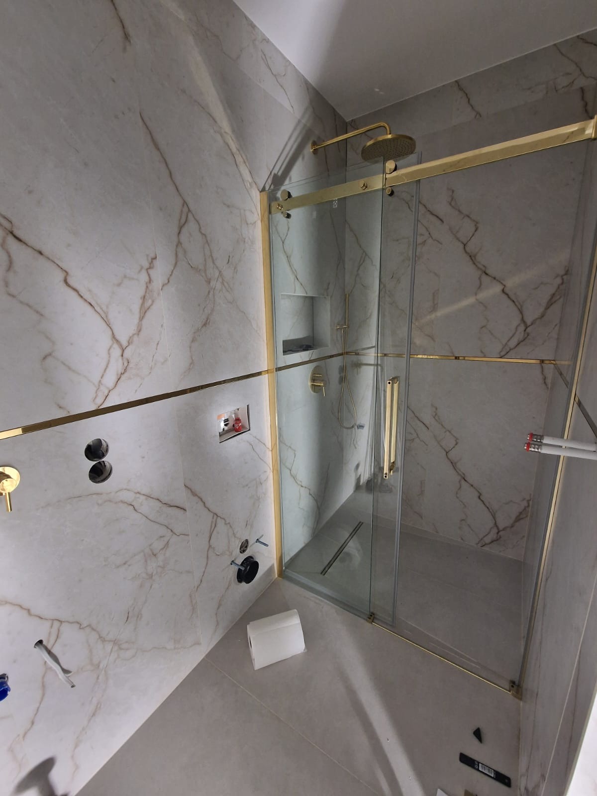 Montaż kabiny prysznicowej z elementami w kolorze złotym na tle marmurowych płytek, widoczne niedokończone prace instalacyjne i rolka papieru na podłodze.