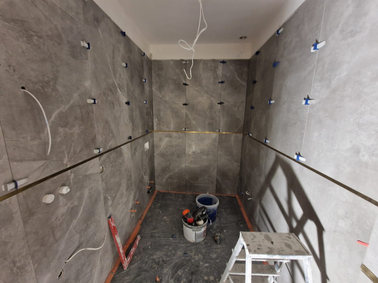 Ściany łazienki w trakcie układania szarych płytek imitujących marmur, z widocznymi krzyżykami dystansowymi i listwami poziomującymi, oraz drabinką i wiadrami z narzędziami na podłodze.