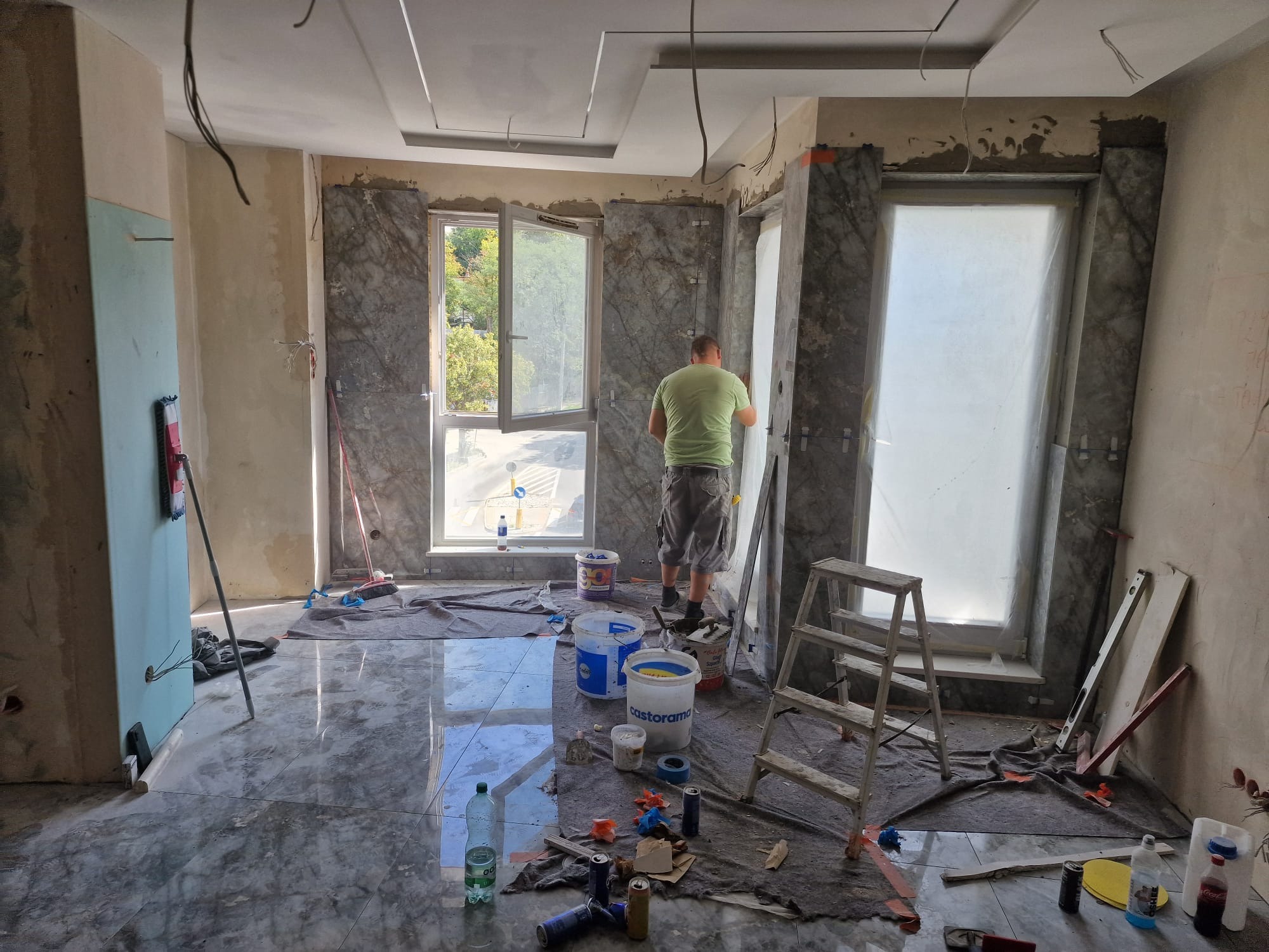 Wnętrze w trakcie prac wykończeniowych, widoczne marmurowe płyty wokół okien, rozłożone narzędzia i drabina, podłoga wyłożona błyszczącymi płytkami imitującymi marmur.