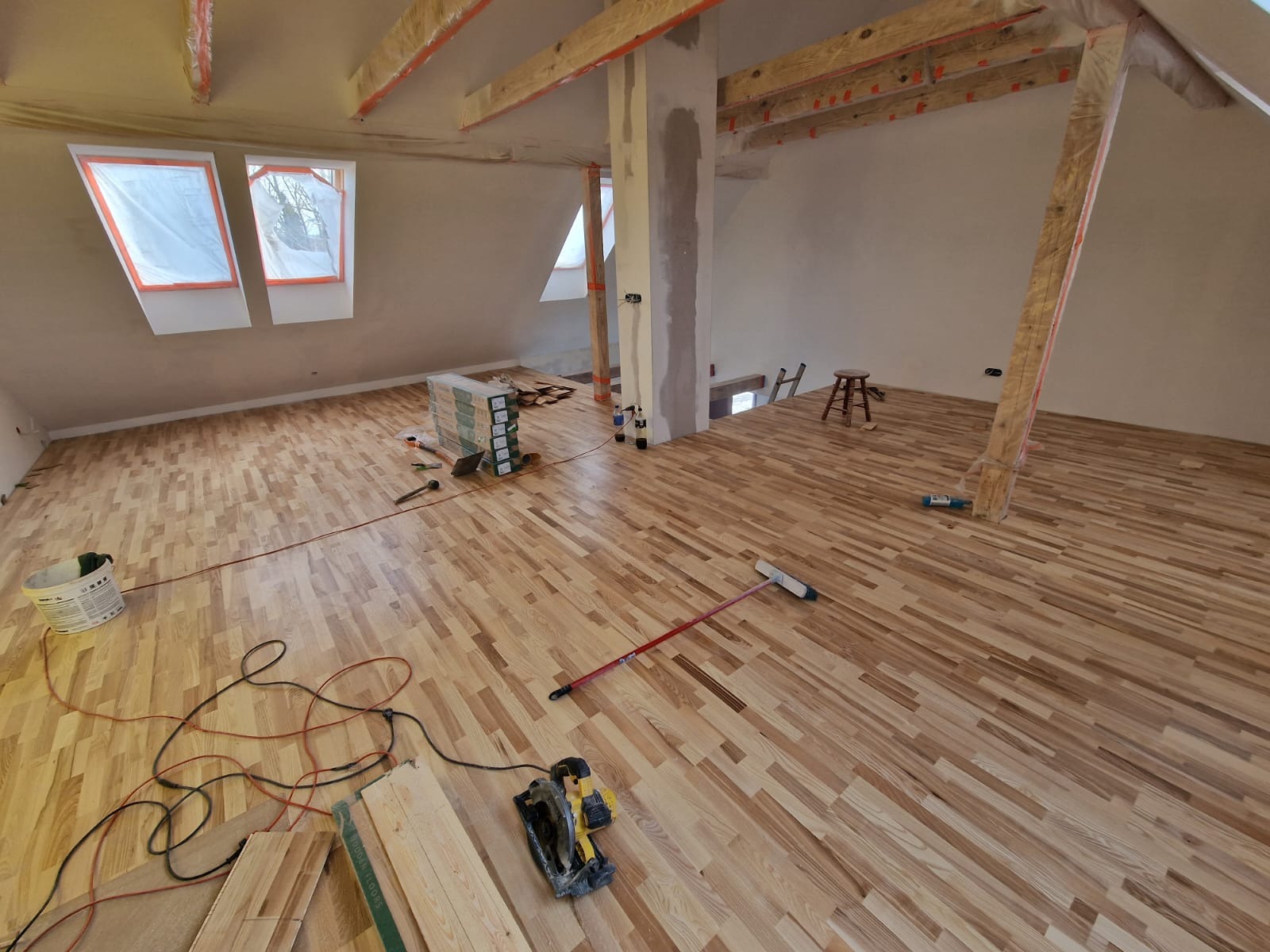Podłoga z parkietu ułożona na poddaszu, widoczne narzędzia pracy, puszka z klejem i paczki z panelami.