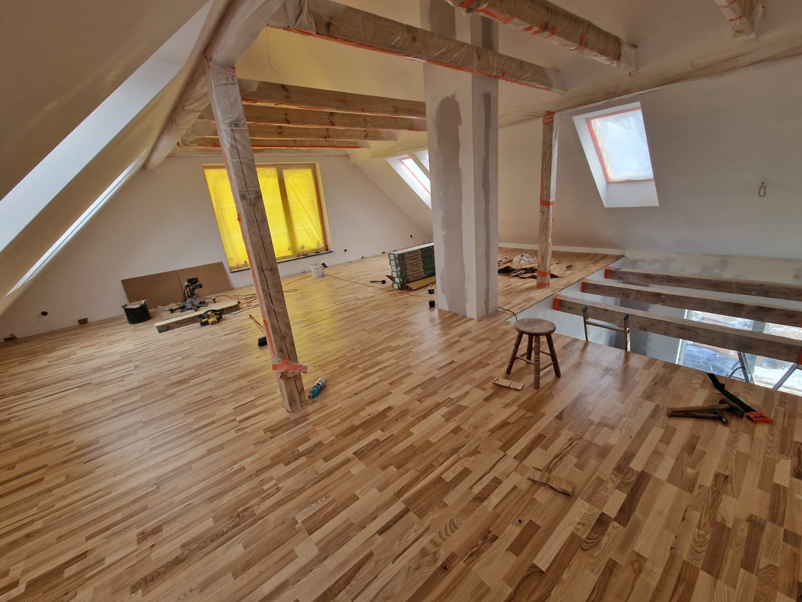 Montaż parkietu w jasnym poddaszu, drewniane belki zabezpieczone folią, widoczne narzędzia i materiały budowlane.
