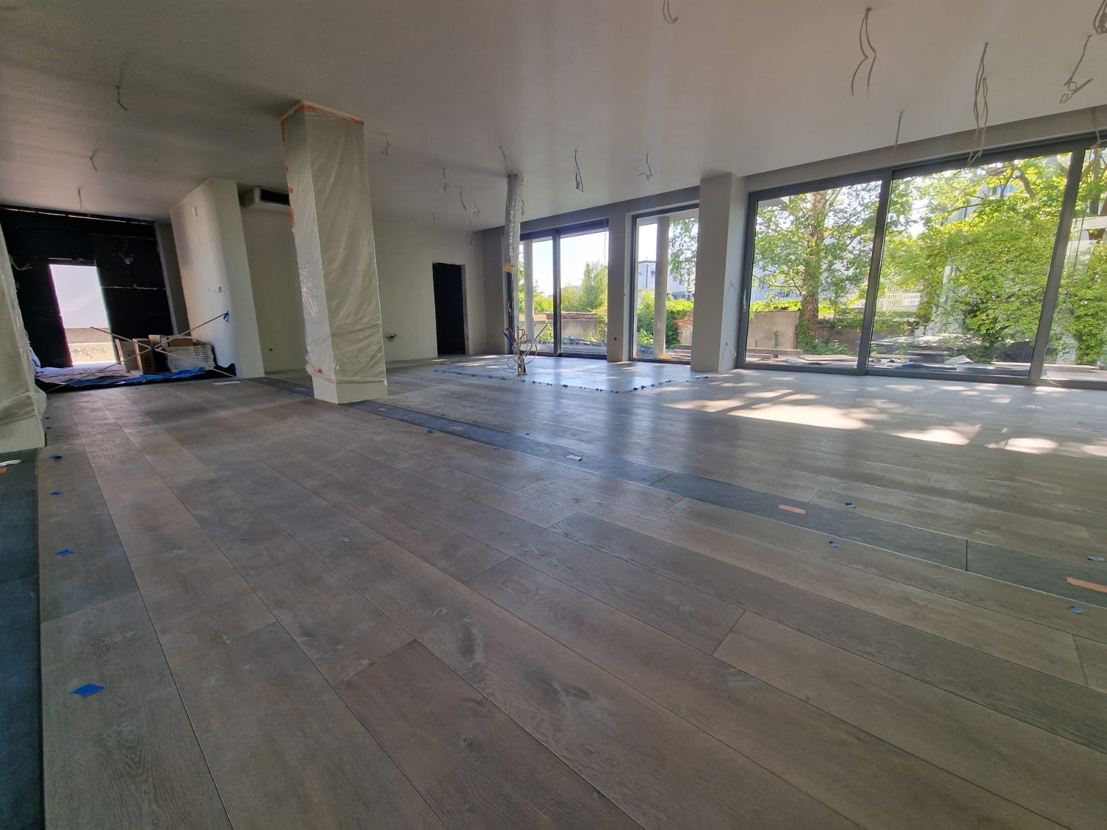 Przestronne wnętrze w trakcie prac wykończeniowych: podłoga z paneli imitujących drewno, duże okna z widokiem na zieleń, zabezpieczone folią filary i przygotowane do instalacji oświetlenie.