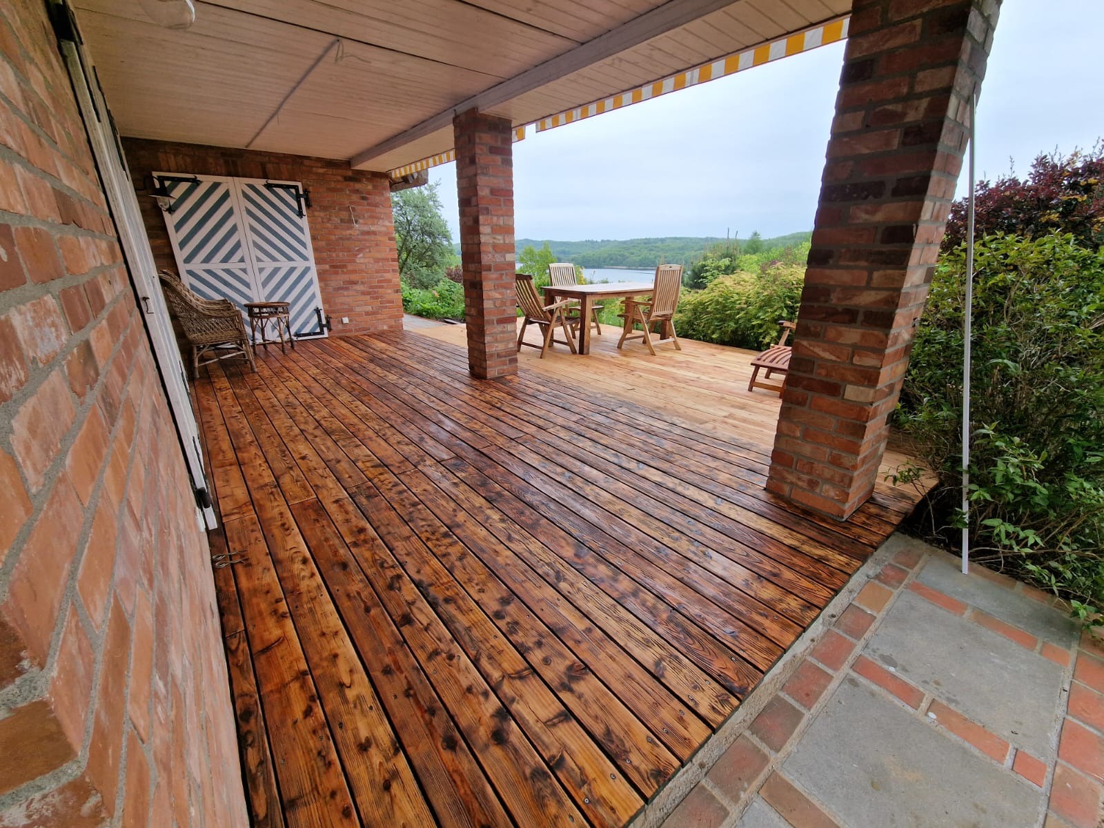 Zadaszony taras z drewnianą podłogą po renowacji, widok na jezioro i zieleń, ceglane filary i białe drzwi z dekoracyjnymi paskami.