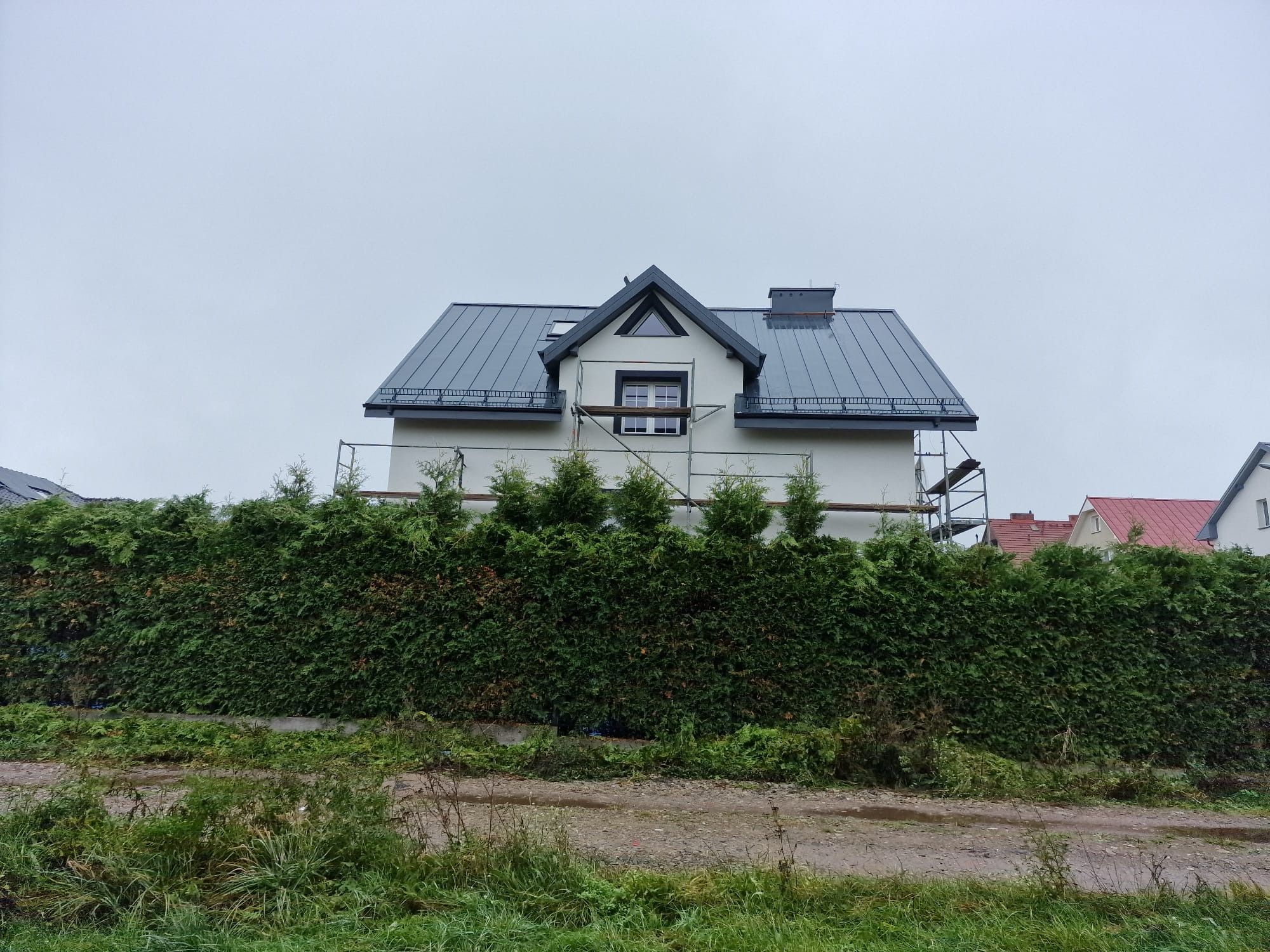 Dom jednorodzinny z szarą blachodachówką, częściowo zasłonięty wysokim żywopłotem, z widocznymi rusztowaniami przy ścianie szczytowej, wskazującymi na prace elewacyjne.