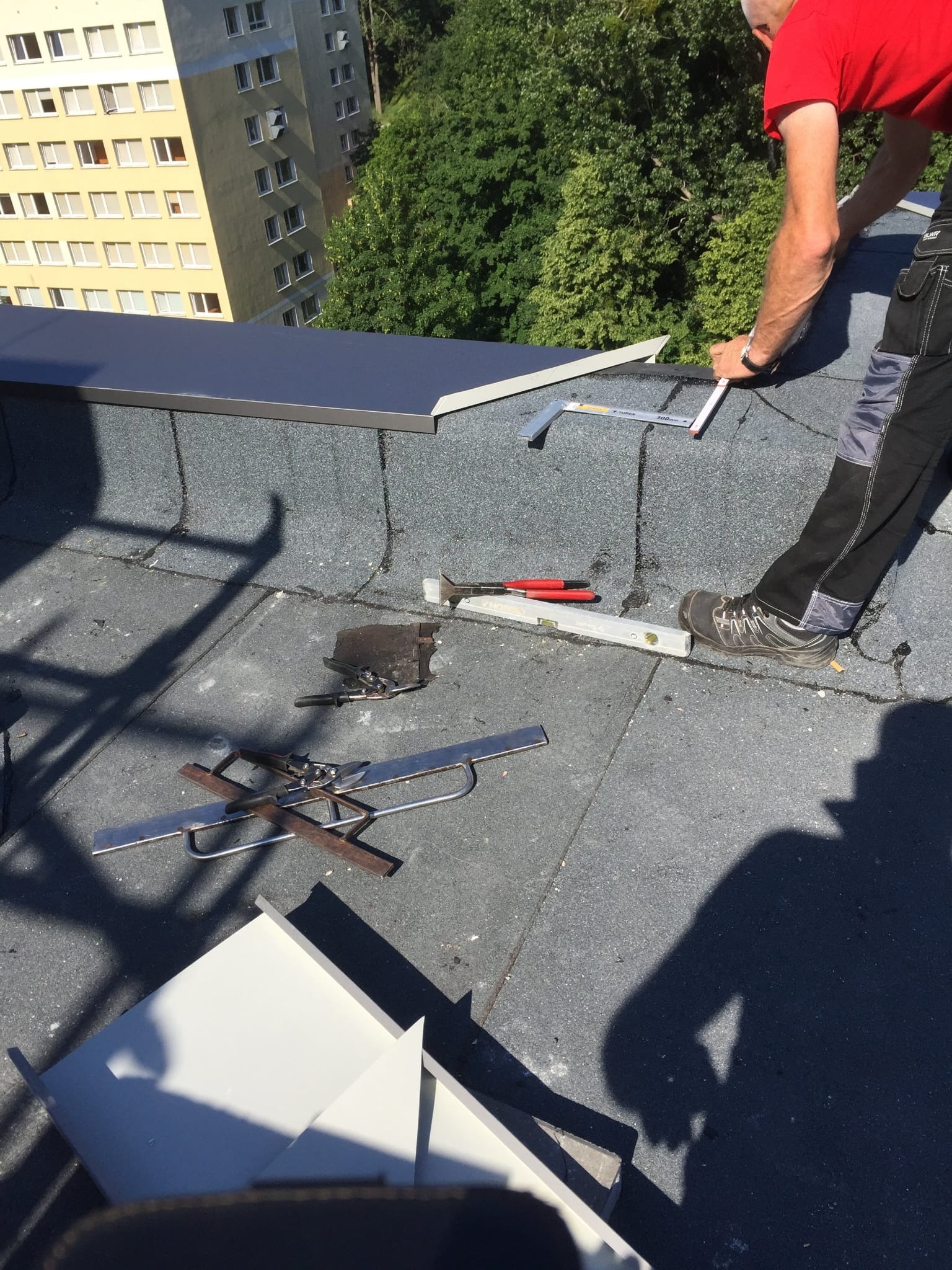 Pracownik w czerwonej koszulce mierzy kąt na dachu pokrytym papą, w tle blok mieszkalny i drzewa, widoczne narzędzia: poziomica, kątownik, nożyce do blachy.