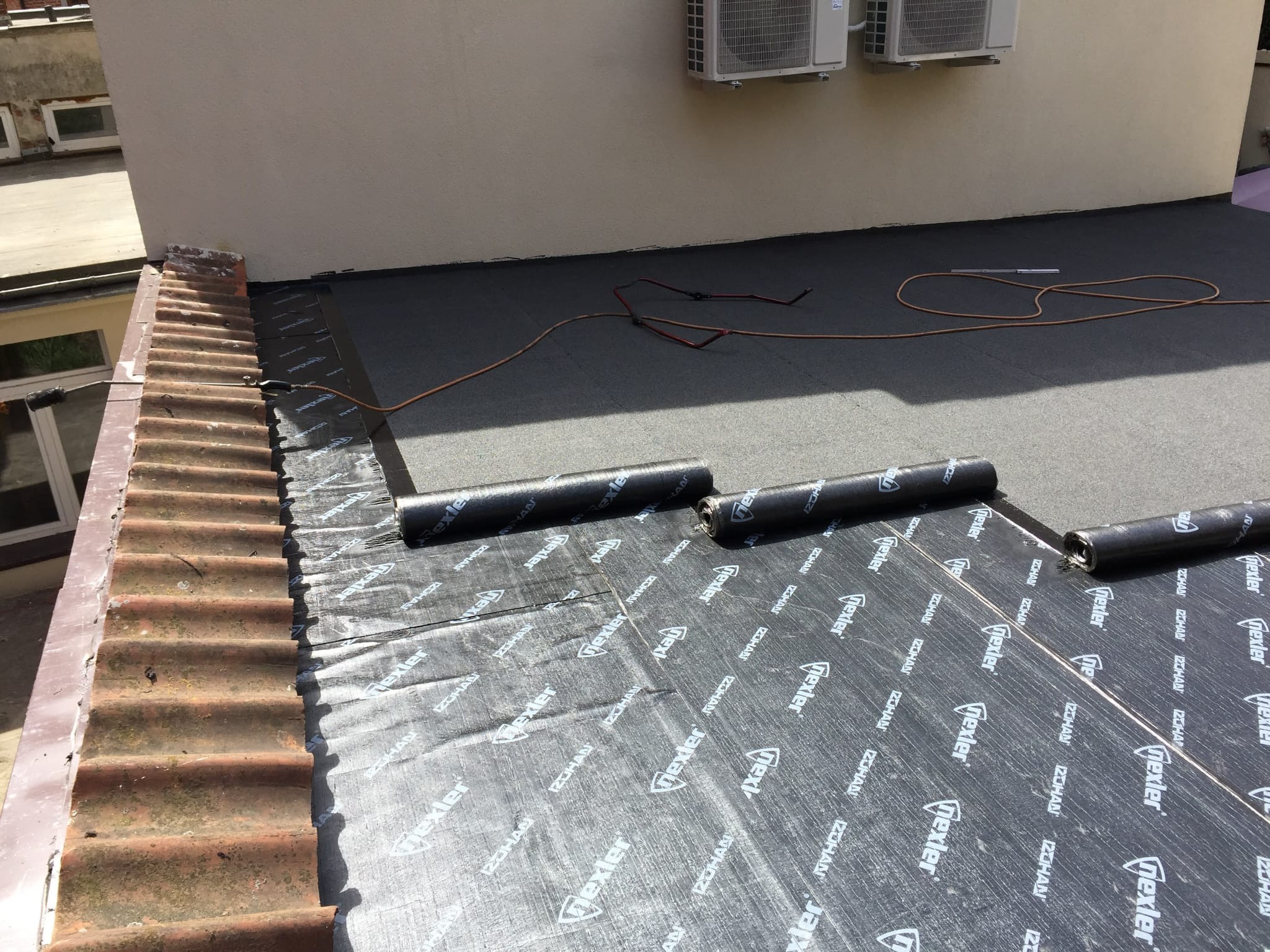 Montaż hydroizolacji na dachu budynku z widocznymi rolkami papy bitumicznej Nexler i palnikiem dekarskim, fragment dachu pokryty starą dachówką