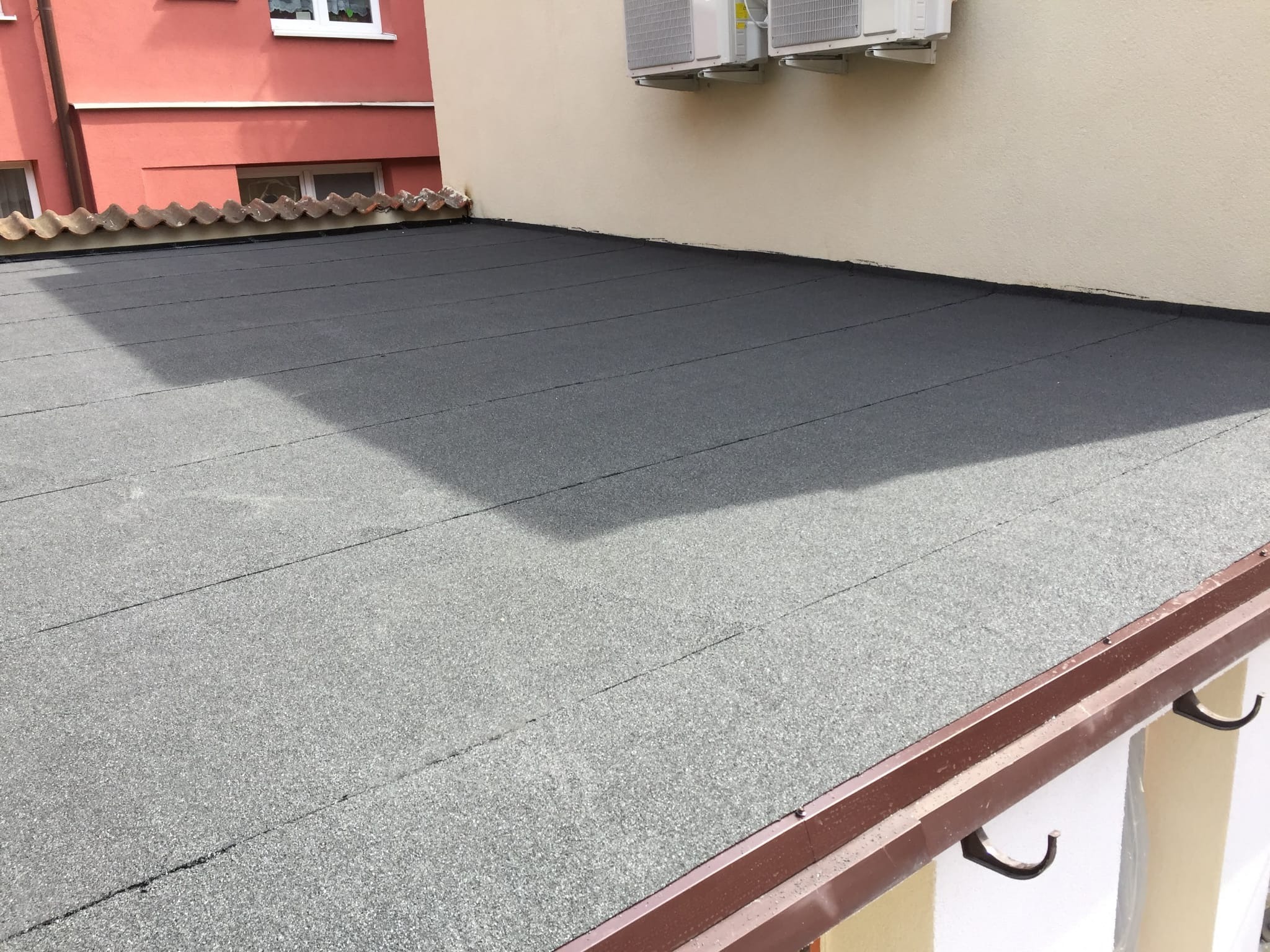 Szczelny, płaski dach pokryty papą termozgrzewalną, z widocznymi liniami łączeń, brązową rynną i fragmentem ściany budynku z klimatyzatorami.