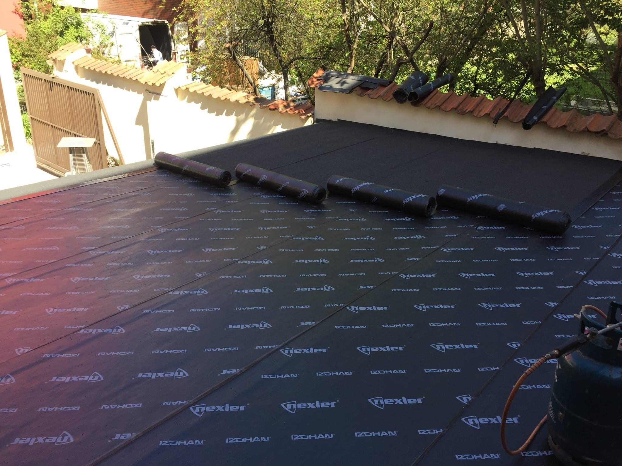 Czarna papa termozgrzewalna z nadrukiem na płaskim dachu, widoczne rolki papy i butla gazowa, w tle mur z dachówką i zieleń.