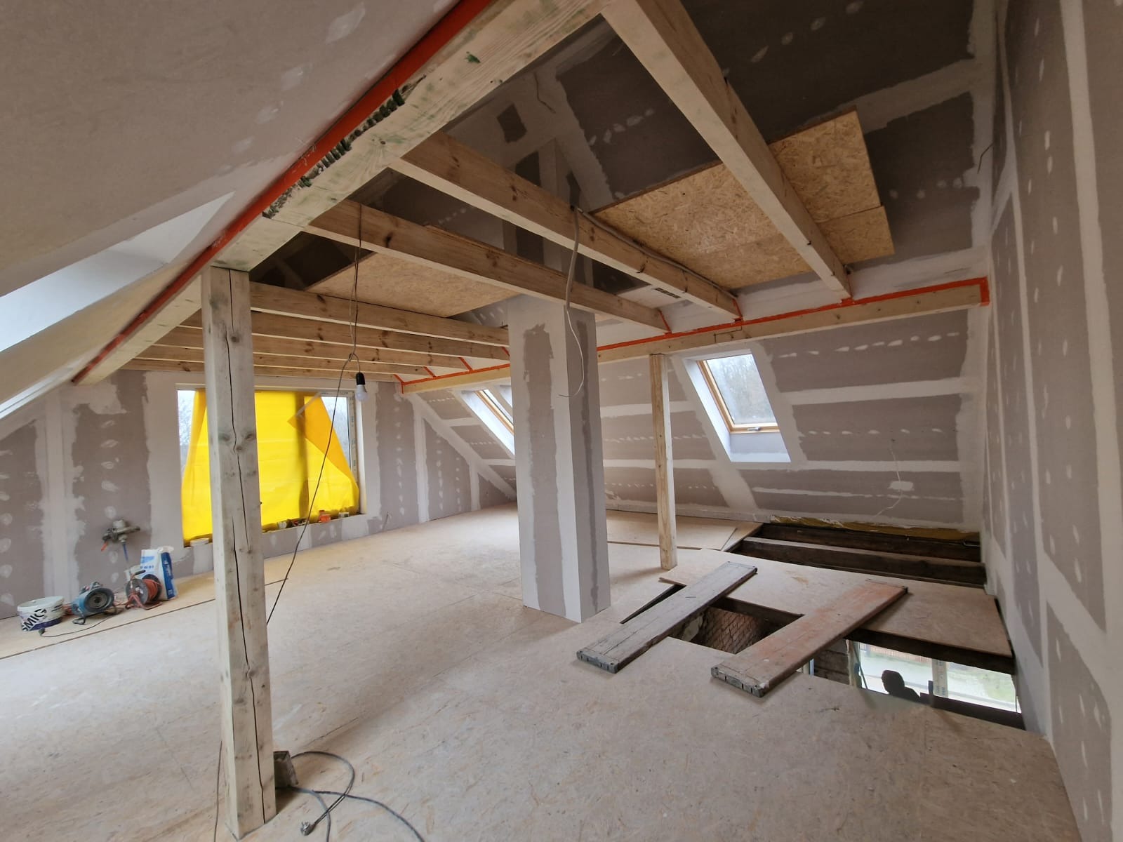Adaptacja poddasza: surowe wnętrze z drewnianą konstrukcją stropu, widoczne płyty OSB, okna dachowe, ściany przygotowane do wykończenia, narzędzia i materiały budowlane w tle.