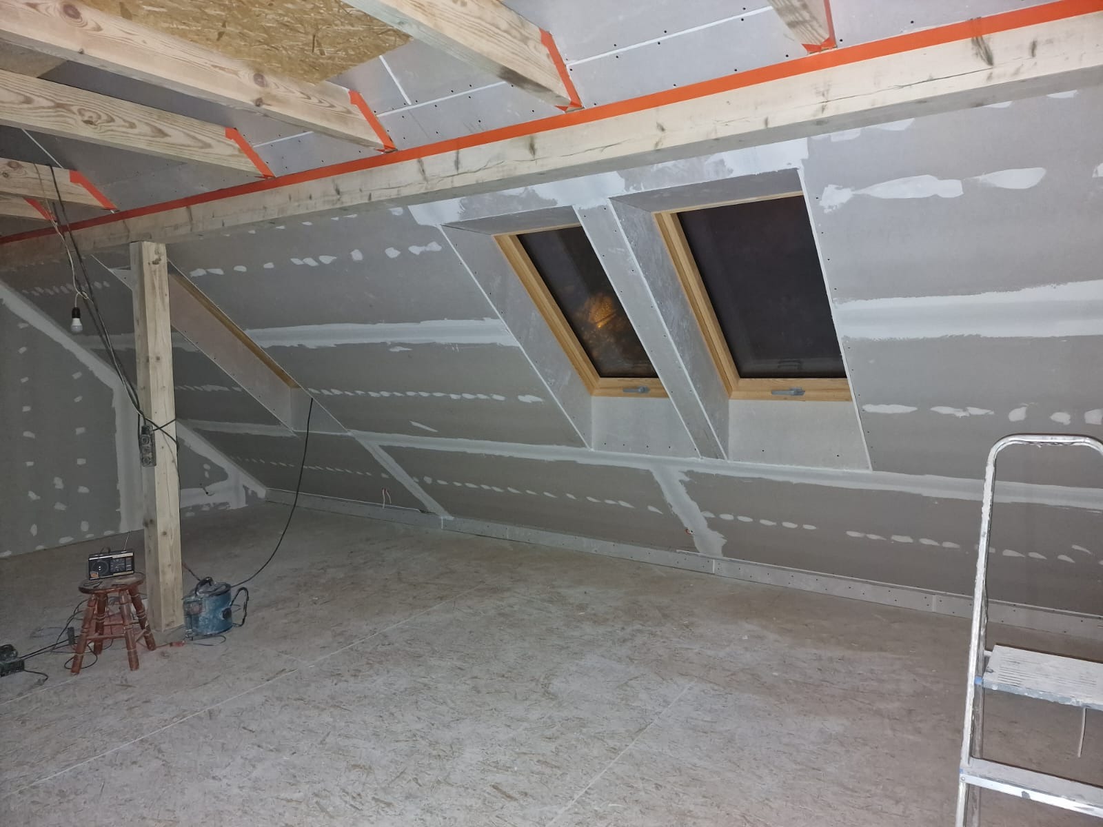 Poddasze w trakcie adaptacji, widoczne płyty gipsowo-kartonowe na ścianach i suficie, drewniana konstrukcja dachu z pomarańczową taśmą, dwa okna dachowe z drewnianymi ramami, podłoga z płyt OSB...