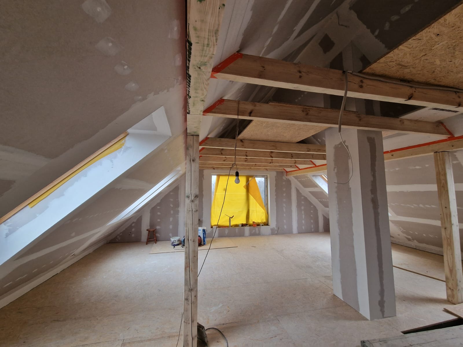 Poddasze w trakcie adaptacji: ściany pokryte płytami gipsowo-kartonowymi, drewniana konstrukcja dachu z widocznymi belkami, okno zasłonięte żółtą folią, widoczne narzędzia i materiały budowlane...
