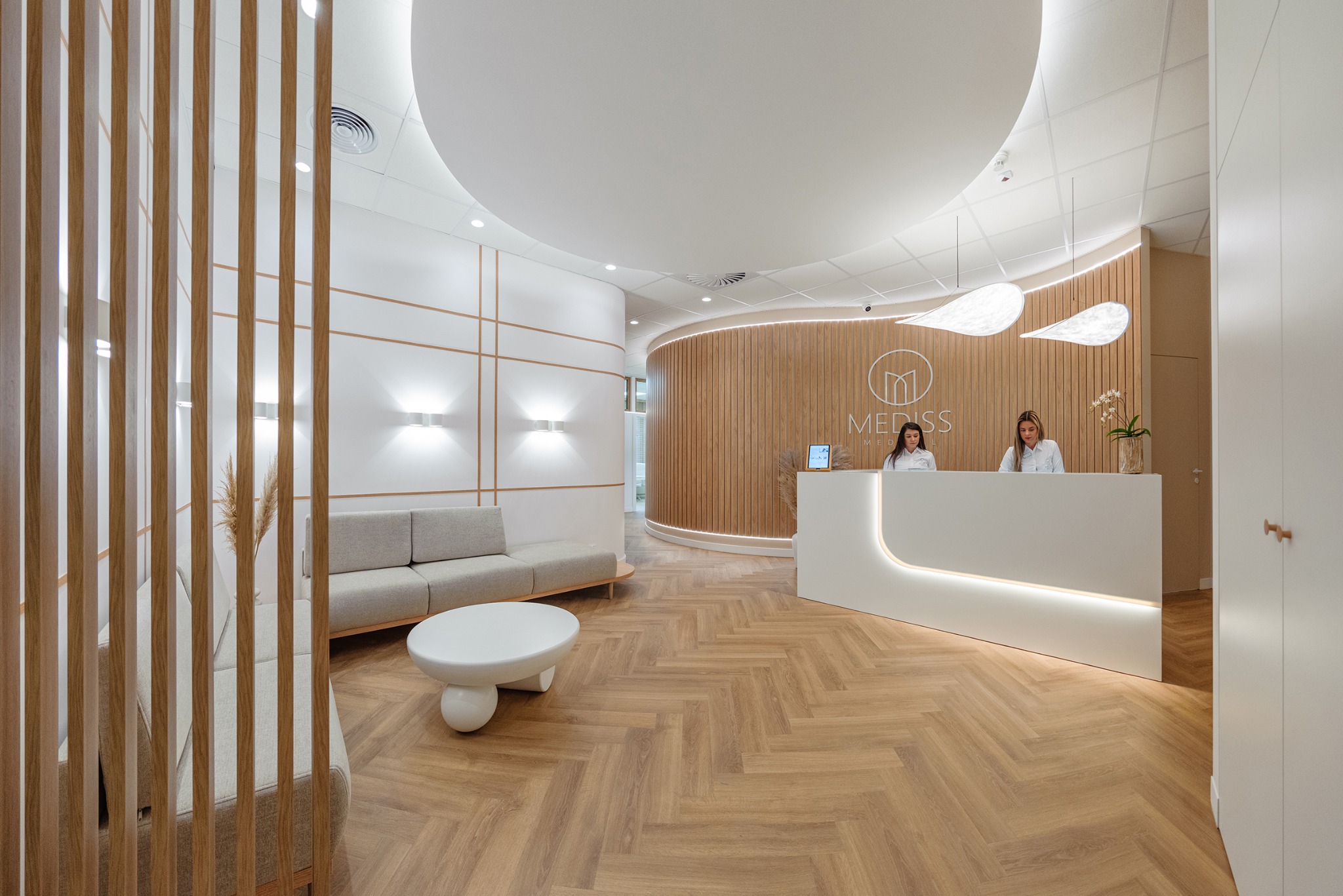 Nowoczesne wnętrze kliniki medycznej z recepcją, drewnianą ścianą, parkietem w jodełkę, minimalistycznym oświetleniem i strefą oczekiwania z szarą sofą i białym stolikiem.