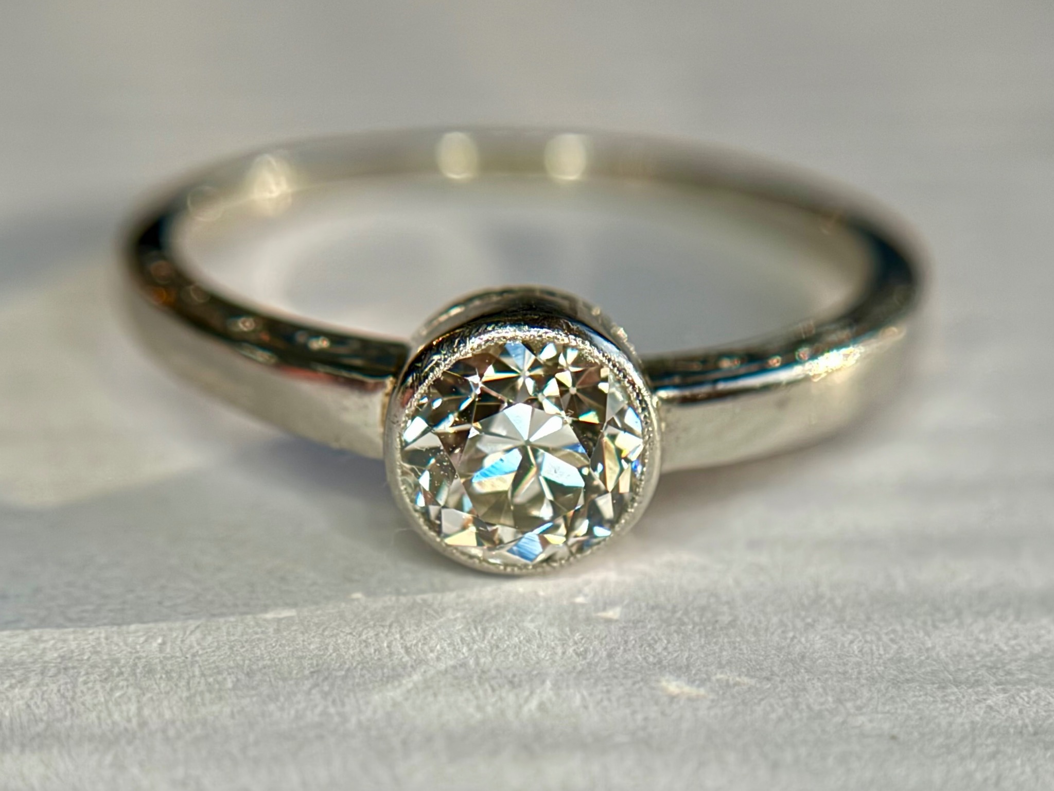 Pierścionek zaręczynowy z okrągłym brylantem w srebrnej oprawie, leżący na białym, teksturowanym tle.