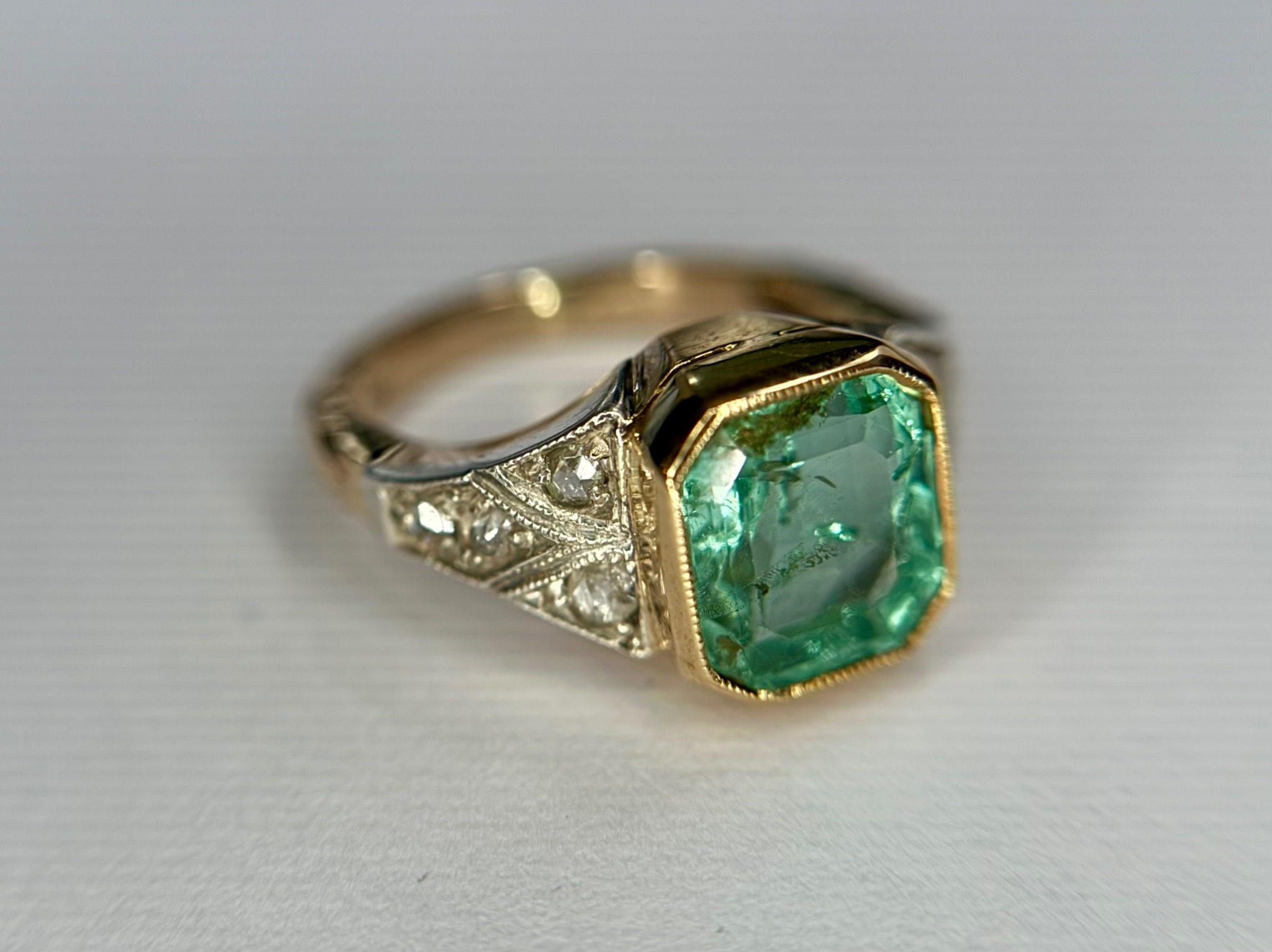 Pierścionek z zielonym szmaragdem o szlifie schodkowym, osadzony w złotej oprawie z dekoracyjnymi elementami z białego złota i brylantami.