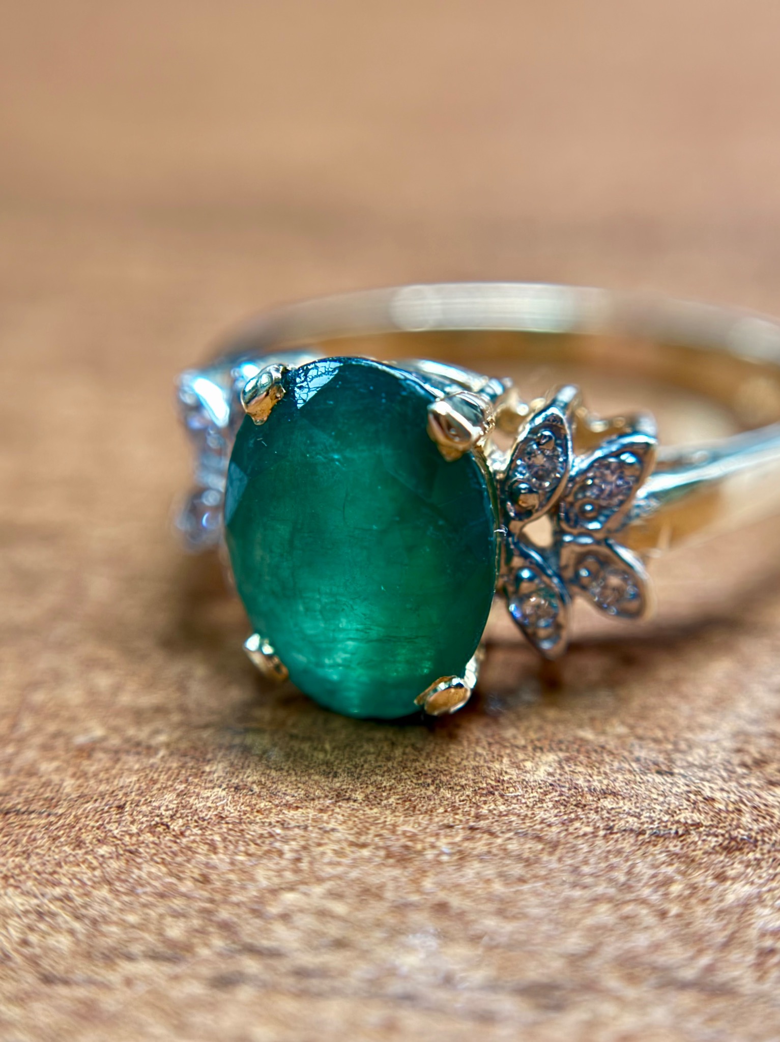 Pierścionek z owalnym szmaragdem i białymi kamieniami w kształcie kwiatów na złotym pierścieniu, leżący na drewnianej powierzchni.