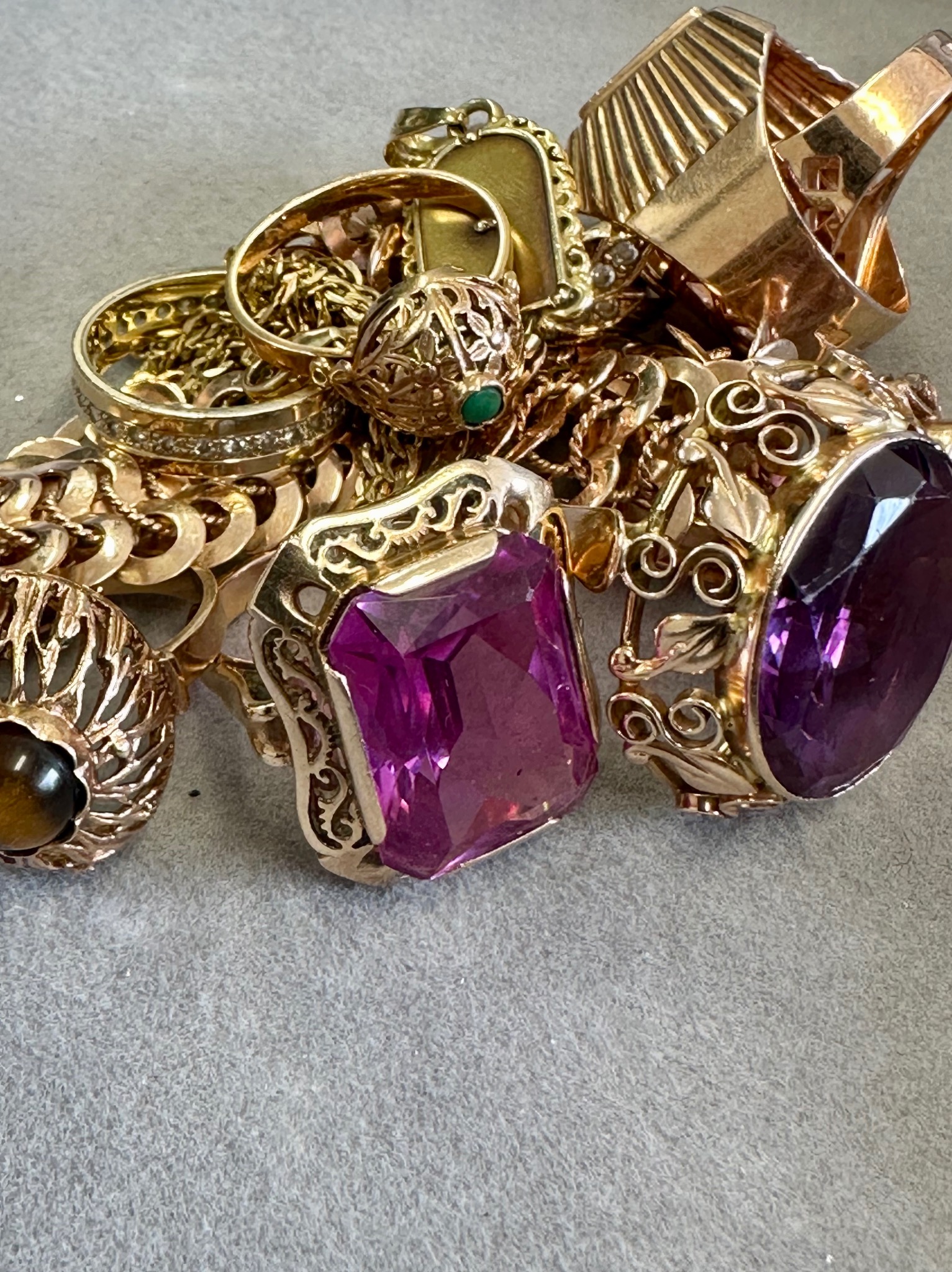 Kolekcja złotej biżuterii z różnymi kamieniami szlachetnymi, w tym pierścionki z ametystem, tygrysim okiem i szmaragdem, ułożona na szarym tle.