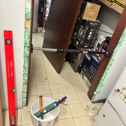 ŁUKASZ GAŁAN - Montaż ościeżnicy drzwiowej z użyciem pianki montażowej i rozpórki. W pobliżu poziomica i pojemnik z narzędziami. W tle widoczne kartony z elektroniką.