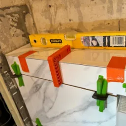 Układanie białych płytek imitujących marmur, poziomowane pomarańczowymi i zielonymi klinami oraz żółtą poziomicą Stanley na tle surowej ściany.