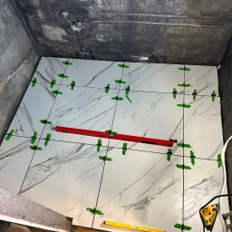 ŁUKASZ GAŁAN - Układanie białych płytek imitujących marmur w łazience, z użyciem poziomicy i klipsów poziomujących. Widoczne surowe ściany i podłoga, etap prac glazurniczych.