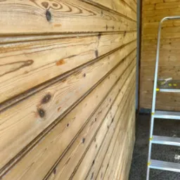 Ściana z desek boazeryjnych o jasnym odcieniu drewna, z widoczną strukturą słojów, oraz fragment drabiny aluminiowej. Ujęcie z boku, podkreślające fakturę drewna.