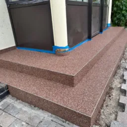 Wykończone schody wejściowe z żywicy kamiennej w odcieniach brązu, zabezpieczone niebieską taśmą malarską przy drzwiach. Fragment kostki brukowej w tle.