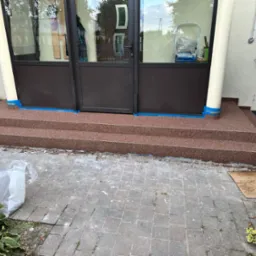 Nowe, brązowe schody wejściowe z żywicy, zabezpieczone niebieską taśmą malarską, prowadzą do ciemnych drzwi. Widoczne rośliny i kostka brukowa.