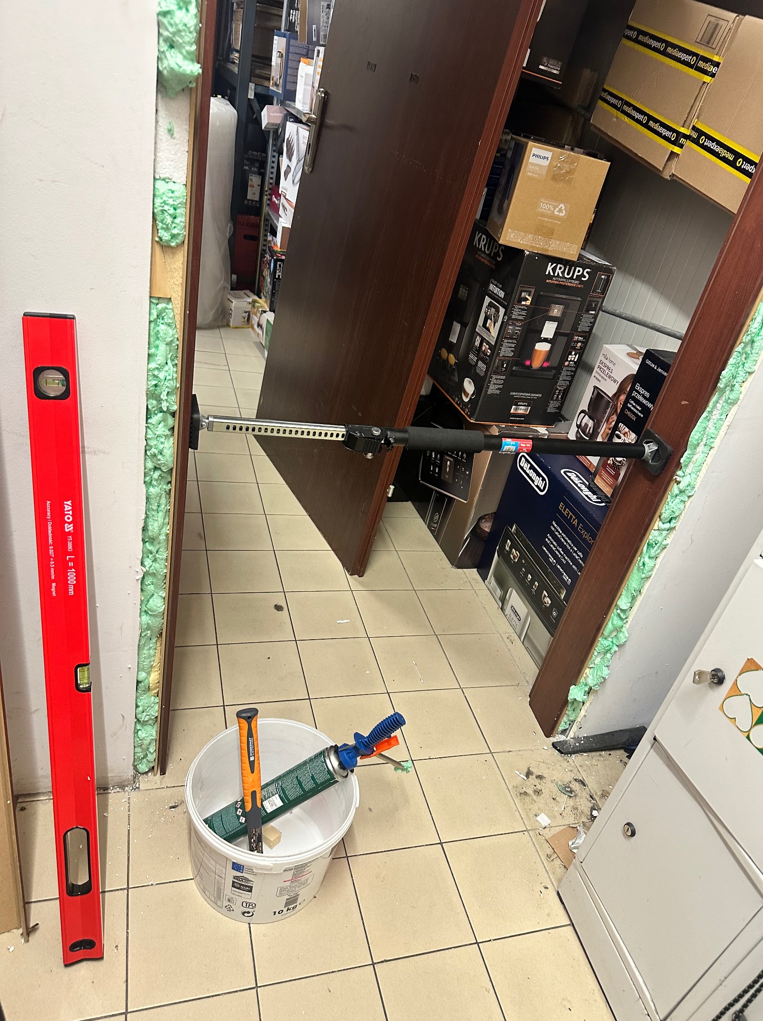 Montaż ościeżnicy drzwiowej z użyciem pianki montażowej i rozpórki. W pobliżu poziomica i pojemnik z narzędziami. W tle widoczne kartony z elektroniką.