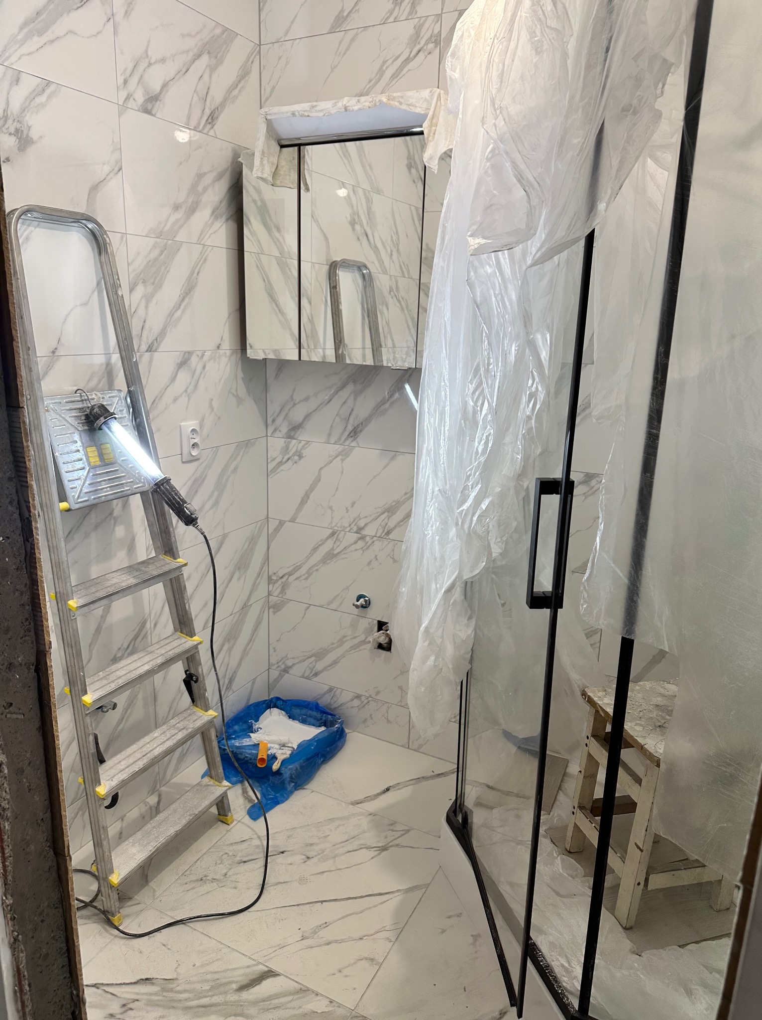 Remont łazienki w trakcie: marmurowe płytki, kabina prysznicowa osłonięta folią, drabina z lampą i narzędzia malarskie. Widoczny etap prac wykończeniowych.