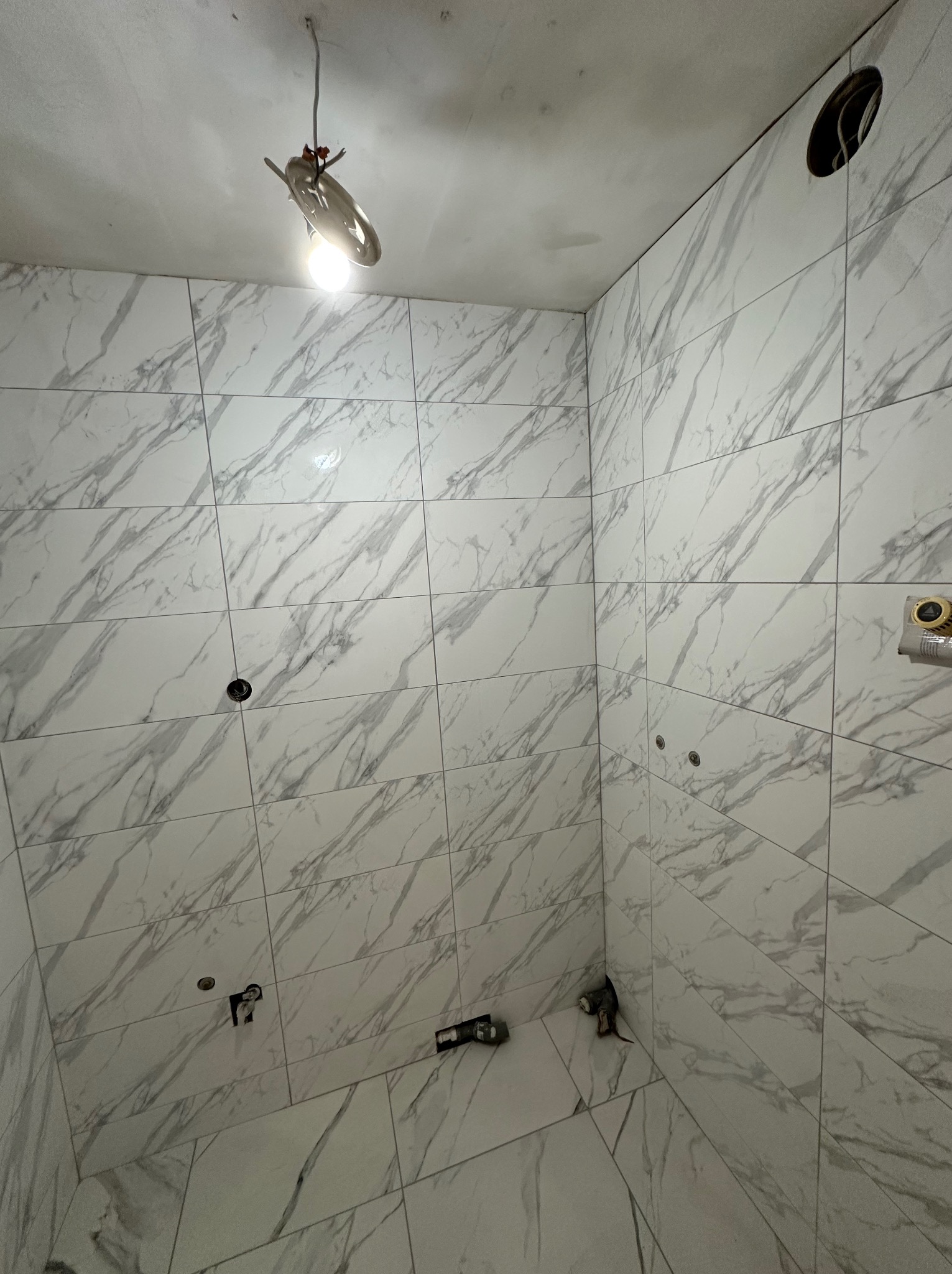 Wnętrze łazienki w trakcie remontu: białe płytki imitujące marmur na ścianach, widoczne otwory na instalacje. U góry prowizoryczne oświetlenie.