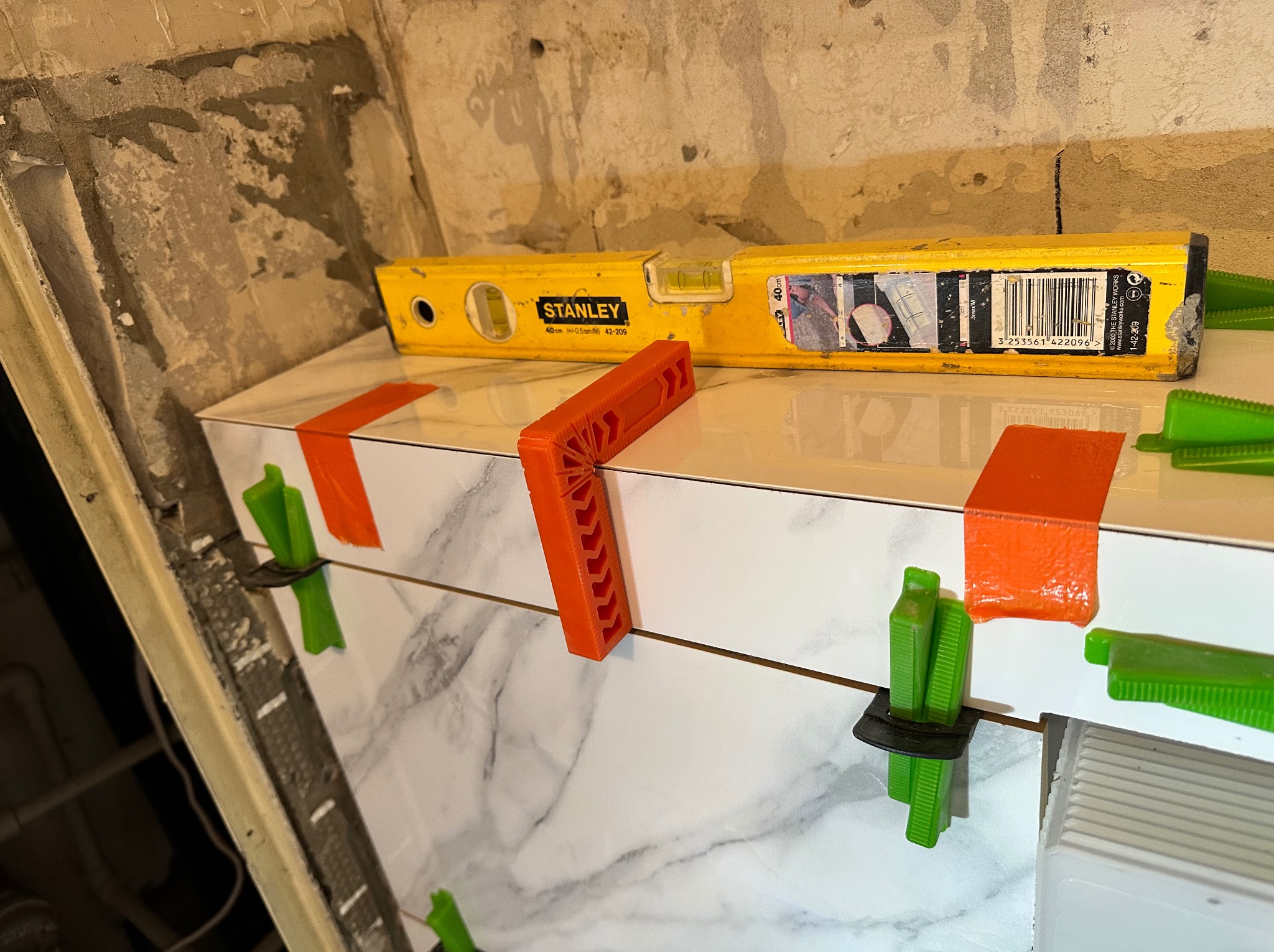 Układanie białych płytek imitujących marmur, poziomowane pomarańczowymi i zielonymi klinami oraz żółtą poziomicą Stanley na tle surowej ściany.