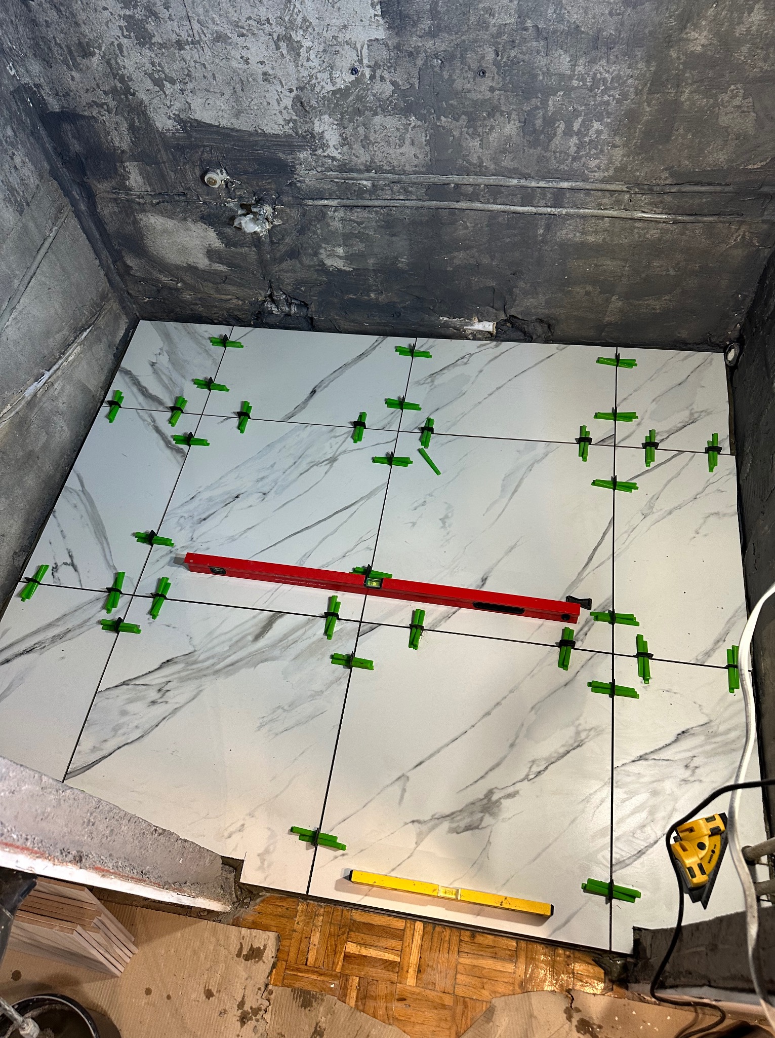 Układanie białych płytek imitujących marmur w łazience, z użyciem poziomicy i klipsów poziomujących. Widoczne surowe ściany i podłoga, etap prac glazurniczych.