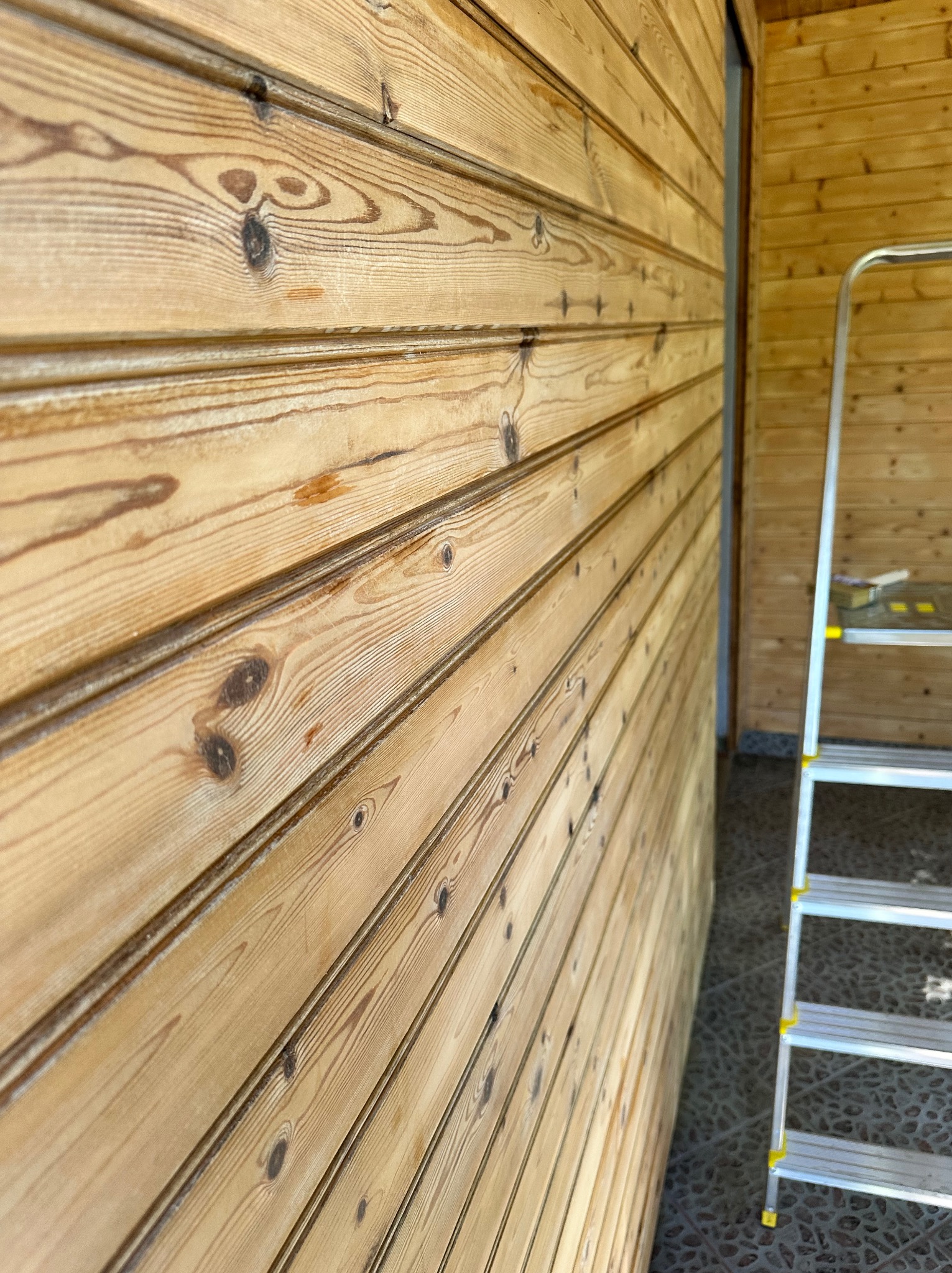 Ściana z desek boazeryjnych o jasnym odcieniu drewna, z widoczną strukturą słojów, oraz fragment drabiny aluminiowej. Ujęcie z boku, podkreślające fakturę drewna.