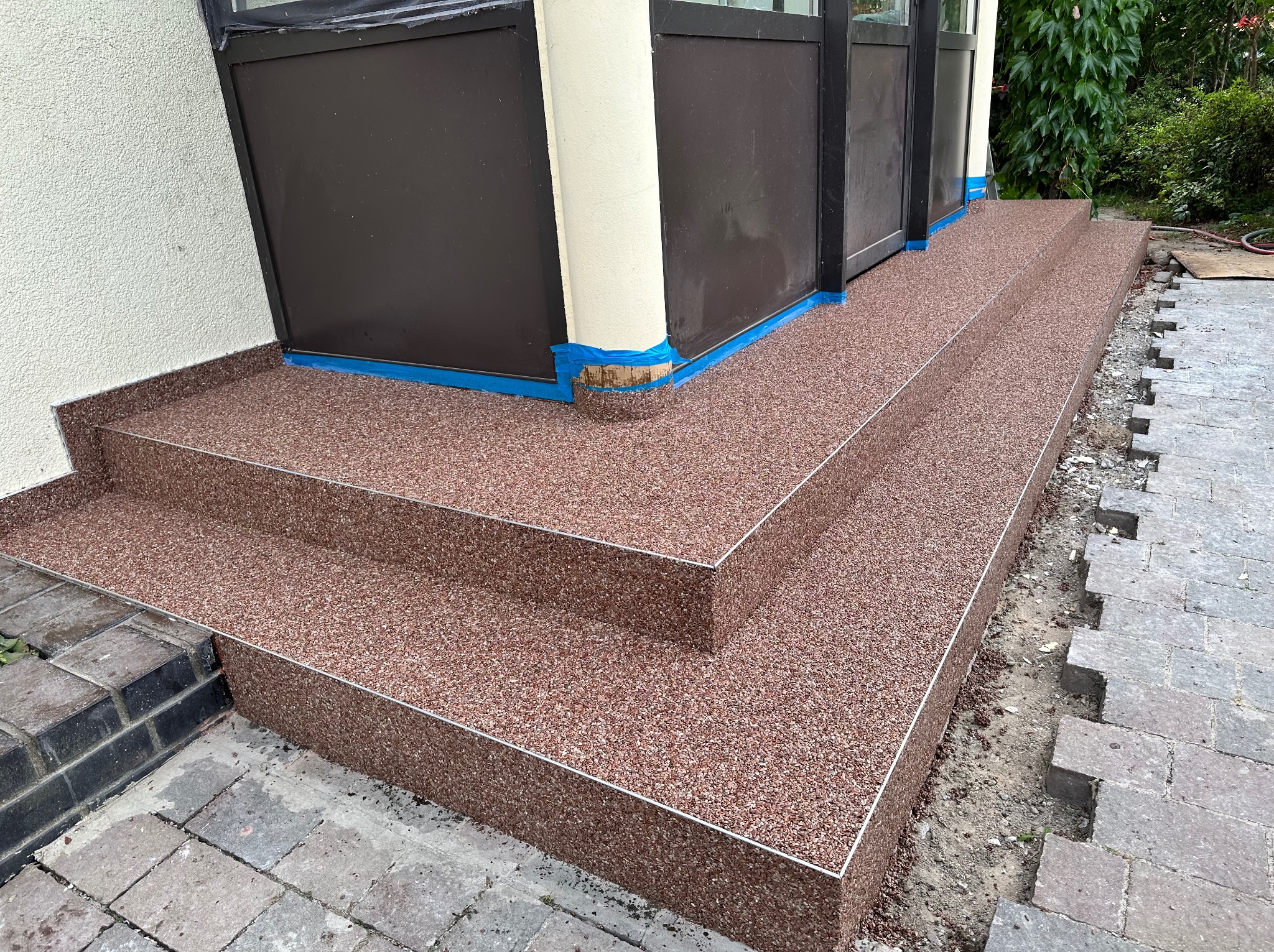 Wykończone schody wejściowe z żywicy kamiennej w odcieniach brązu, zabezpieczone niebieską taśmą malarską przy drzwiach. Fragment kostki brukowej w tle.
