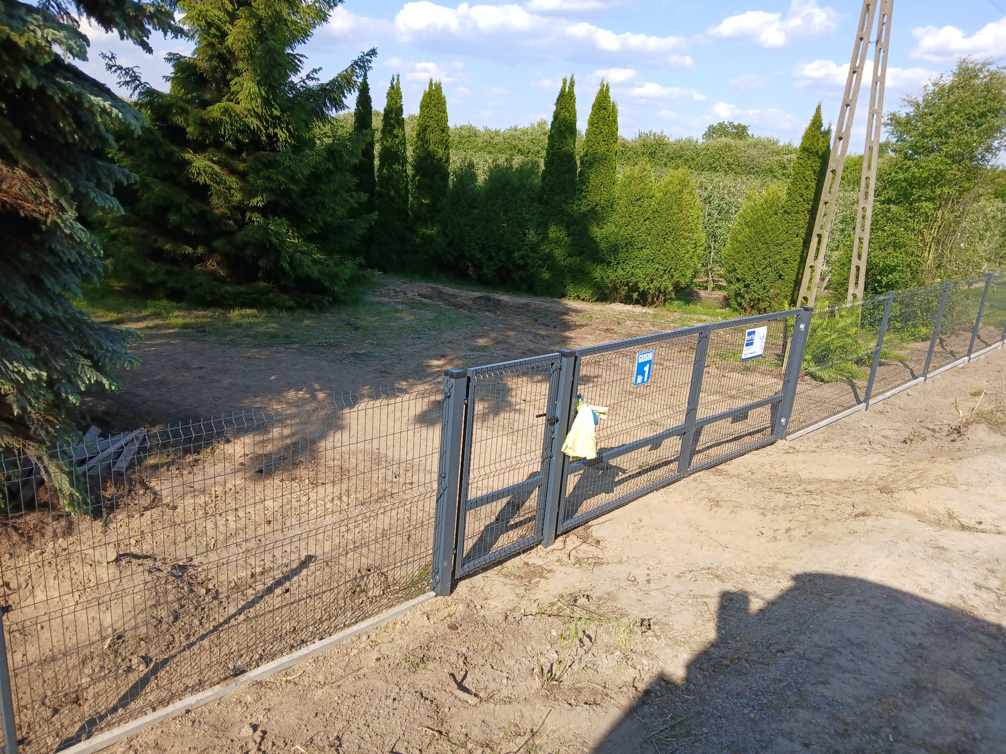 Szare, metalowe ogrodzenie panelowe z bramką na tle zieleni i nieba. Na bramce wisi żółta torba. Widoczne słupy energetyczne i cień osoby.