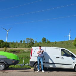 JEST ELEGANCKO MYCIE CIŚNIENIOWE KAROL GOLUBEK - Pracownik z lancą ciśnieniową stoi obok przyczepy z myjką JMB-S+ 200/18 i białego vana na tle zielonego wzgórza z turbinami wiatrowymi.