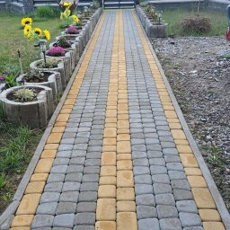 JEST ELEGANCKO MYCIE CIŚNIENIOWE KAROL GOLUBEK - Ścieżka z kostki brukowej w odcieniach szarości i żółci prowadząca do bramy. Po bokach rabaty z kwiatami, w tle drzewa.
