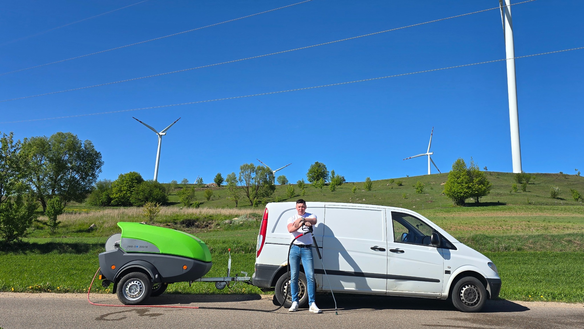 Pracownik z lancą ciśnieniową stoi obok przyczepy z myjką JMB-S+ 200/18 i białego vana na tle zielonego wzgórza z turbinami wiatrowymi.