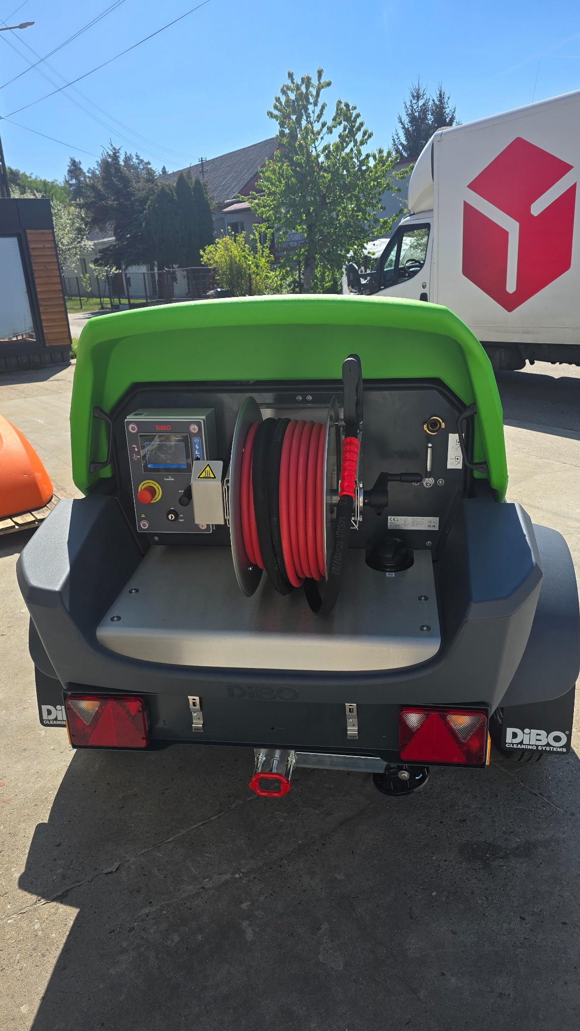 Profesjonalna maszyna do czyszczenia Dibo z nawiniętym czerwonym wężem, widok z tyłu, na tle budynku i samochodu dostawczego w słoneczny dzień.