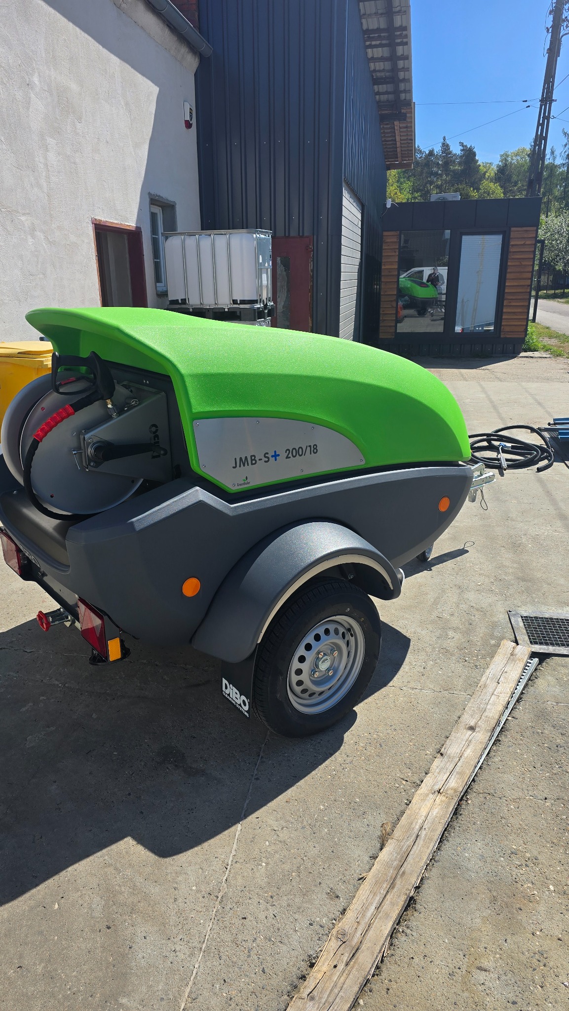 Przyczepa z myjką ciśnieniową Dibo JMB-S+ 200/18 na betonowym podłożu, w tle budynki. Zielono-szara maszyna gotowa do użycia.