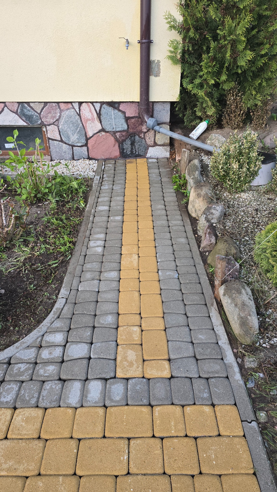 Ścieżka z kostki brukowej w odcieniach szarości i żółci prowadząca wzdłuż ściany budynku z elementami kamiennymi. Widoczny fragment rynny, roślinność i kamienie ozdobne.