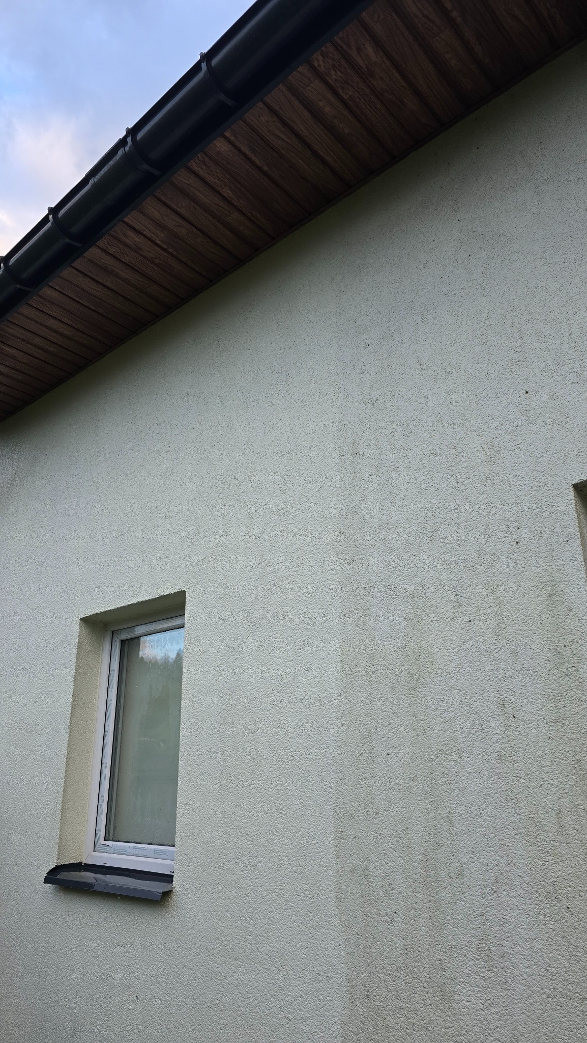 Ściana budynku z widocznymi śladami zabrudzeń i zacieków, okno z białą ramą, brązowe podbitki dachowe i czarna rynna. Widoczne efekty atmosferyczne na elewacji.
