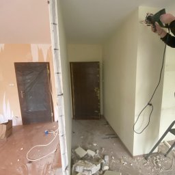 ANDRIY ANDRIISHYN - Burzenie ściany działowej w mieszkaniu podczas remontu. Widoczne gruz i narzędzie w użyciu, drzwi zabezpieczone folią. Prace wykończeniowe w trakcie.