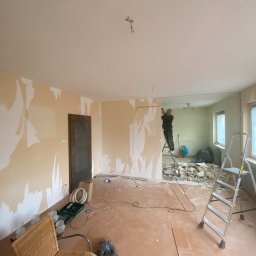 ANDRIY ANDRIISHYN - Demontaż ściany działowej w trakcie remontu. Widoczne narzędzia, drabina, resztki tapet na ścianach i gruz. Osoba na drabinie w głębi kadru.