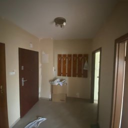 ANDRIY ANDRIISHYN - Korytarz w trakcie remontu: ściany przygotowane do malowania, widoczne ślady szpachlowania, karton i folia malarska, drewniany wieszak na ścianie.