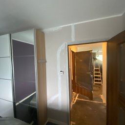 ANDRIY ANDRIISHYN - Ściana po szpachlowaniu z widocznymi śladami łączeń płyt, drzwi zabezpieczone folią malarską. W tle drabina malarska.