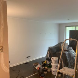 ANDRIY ANDRIISHYN - Pokój po szpachlowaniu, przygotowany do malowania. Widoczne puszki z farbą, drabina, zabezpieczona folią podłoga i gładkie, białe ściany. W tle okno.