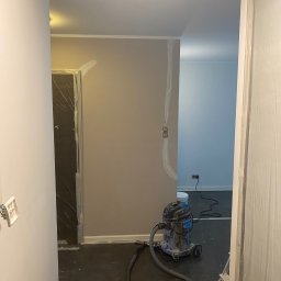 ANDRIY ANDRIISHYN - Przygotowania do malowania: zabezpieczone folią drzwi, ściany po szpachlowaniu, odkurzacz przemysłowy i widoczne puszki elektryczne.